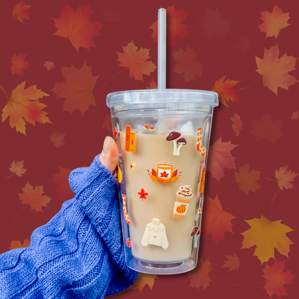 A stylish cold cup featuring autumnal-themed designs such as pumpkins, sweaters, and candles, held by a hand in a cosy blue sweater. Perfect for autumn drinks like iced lattes or pumpkin spice beverages.