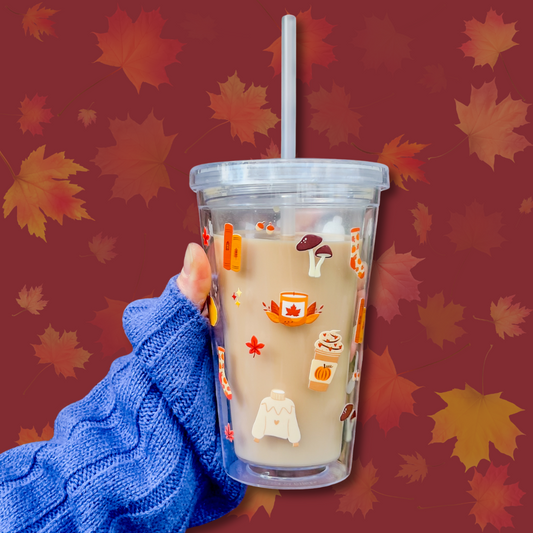 A stylish cold cup featuring autumnal-themed designs such as pumpkins, sweaters, and candles, held by a hand in a cosy blue sweater. Perfect for autumn drinks like iced lattes or pumpkin spice beverages.